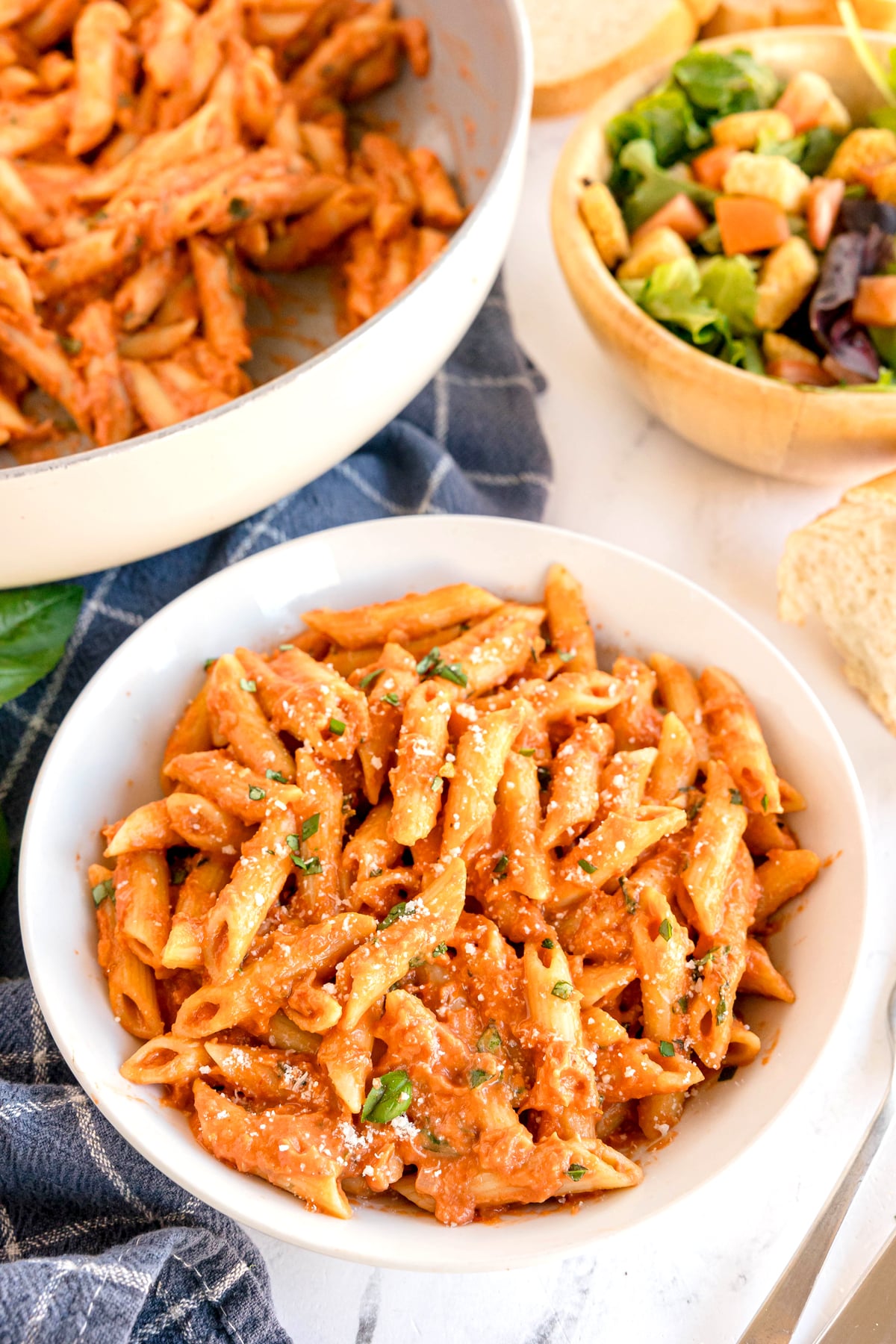 A bowl of Pasta Alla Vodka with tomato sauce and grated cheese, next to a skillet of pasta, a bowl of salad, bread slices, and a blue napkin.