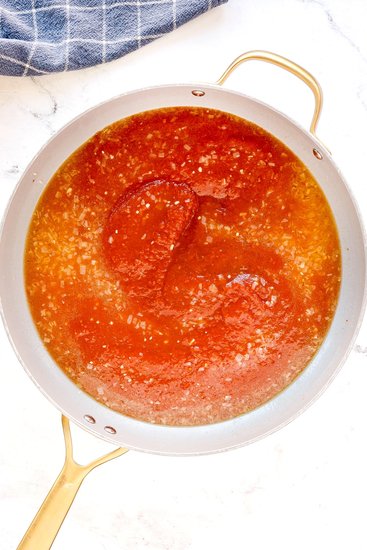 A pan filled with tomato sauce, chicken stock, and sautéed onions on a white surface, for Pasta Alla Vodka recipe, with a blue checkered cloth partially visible in the corner.