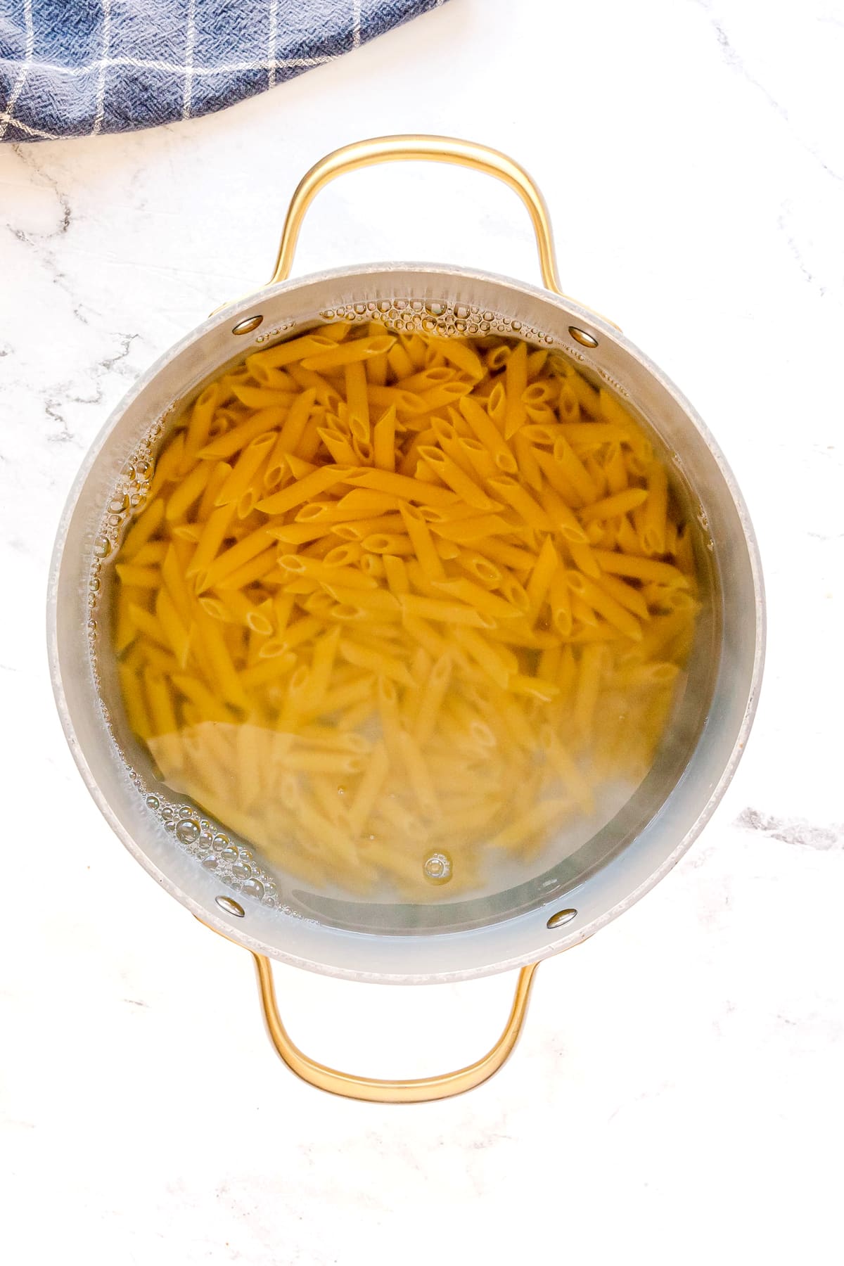 A pot filled with uncooked penne pasta for Pasta Alla Vodka recipe, submerged in water, viewed from above on a white surface.