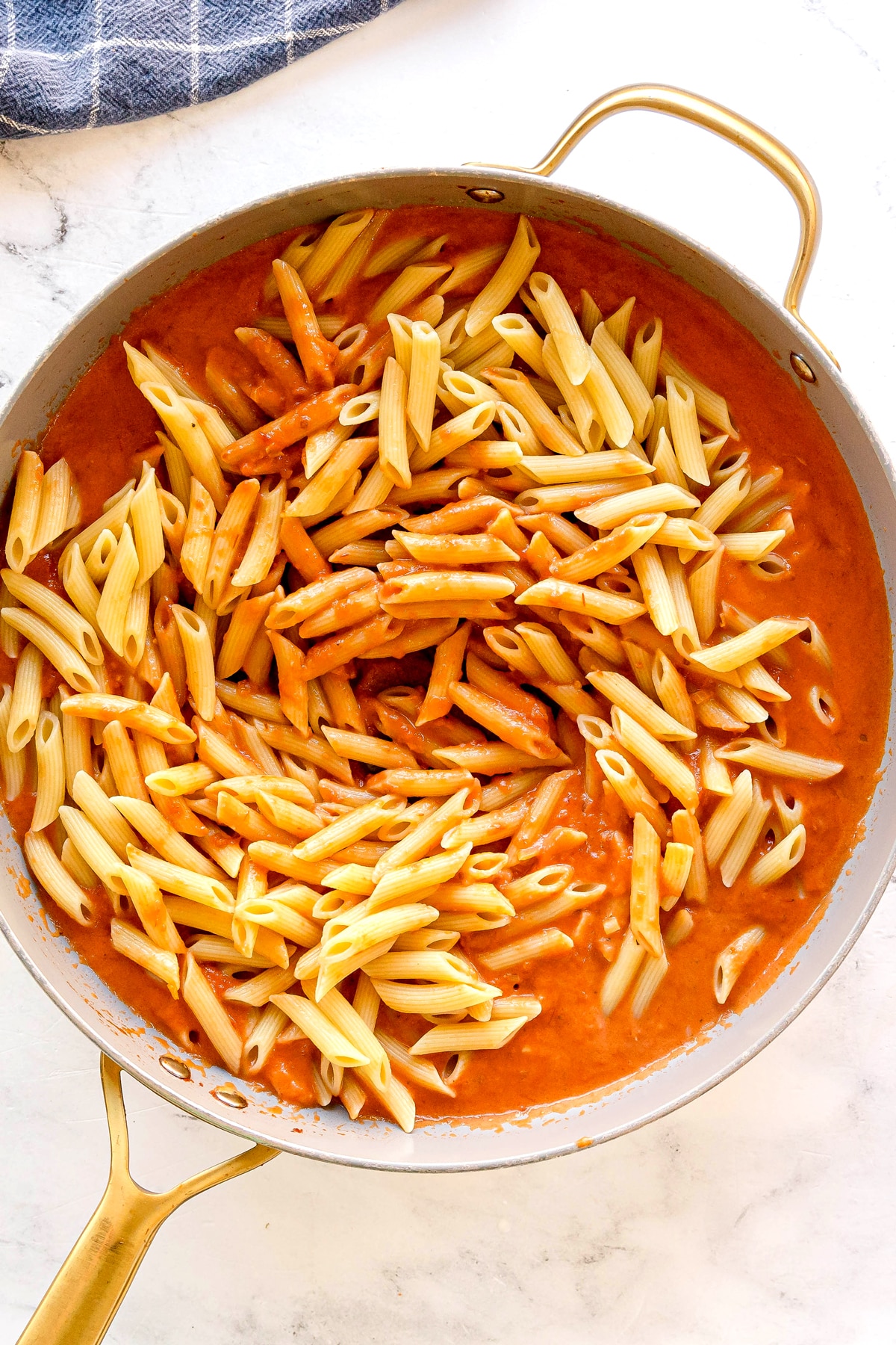 A pan filled with penne pasta partially mixed into a creamy tomato sauce, sitting on a white surface for Pasta Alla Vodka recipe.