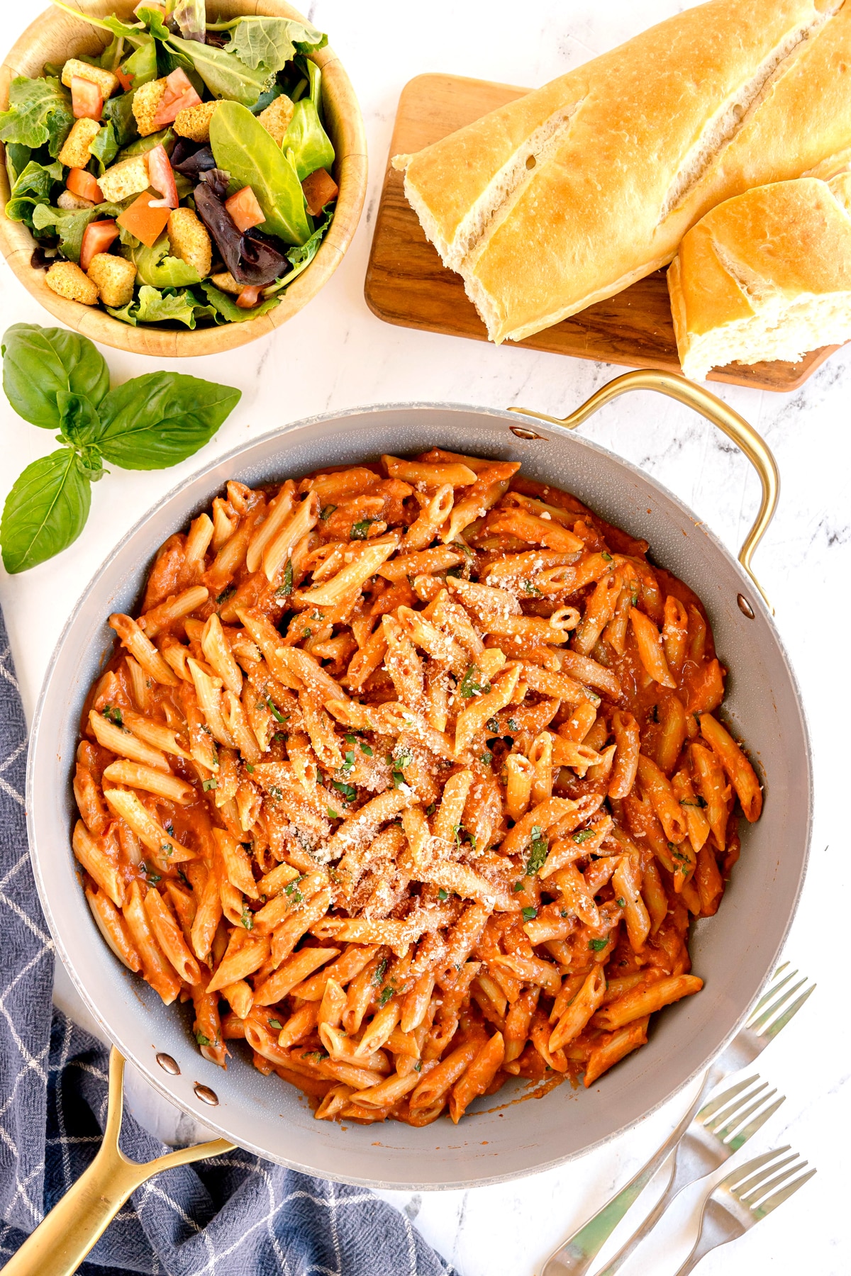 A skillet of Pasta Alla Vodka with red sauce and grated cheese, served with a green salad in a bowl and sliced baguette on a wooden board.