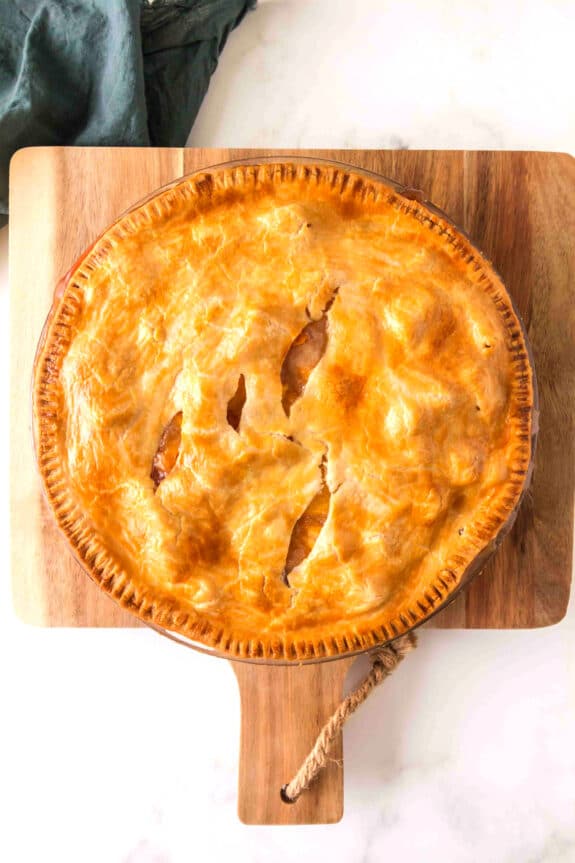 A golden-brown Peach Pie with a flaky crust sits on a wooden cutting board, viewed from above.