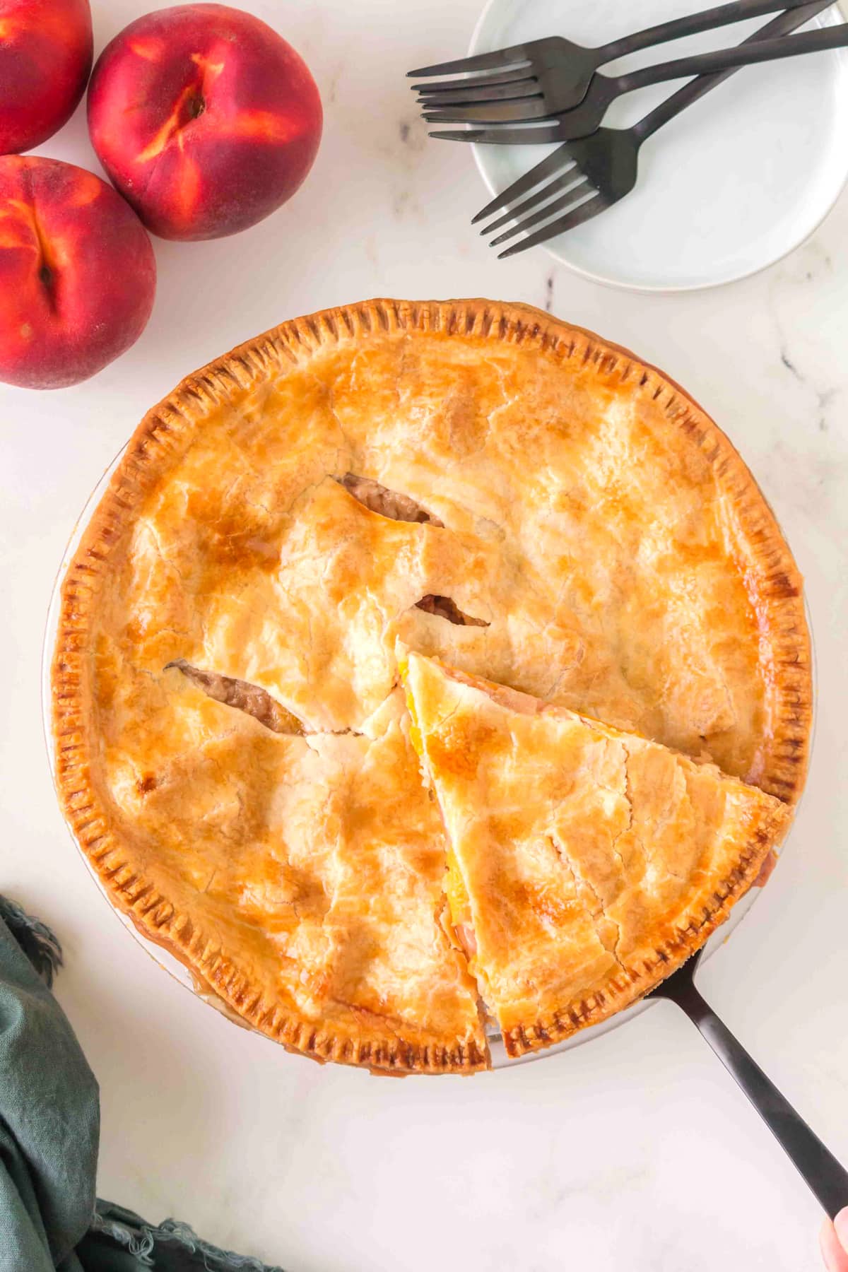 Whole Peach Pie with a golden crust, one slice being served, next to fresh peaches, black forks, and a white plate on a marble surface.