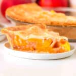 A slice of Peach Pie with a golden, flaky crust sits on a white plate, with the rest of the pie in the background.