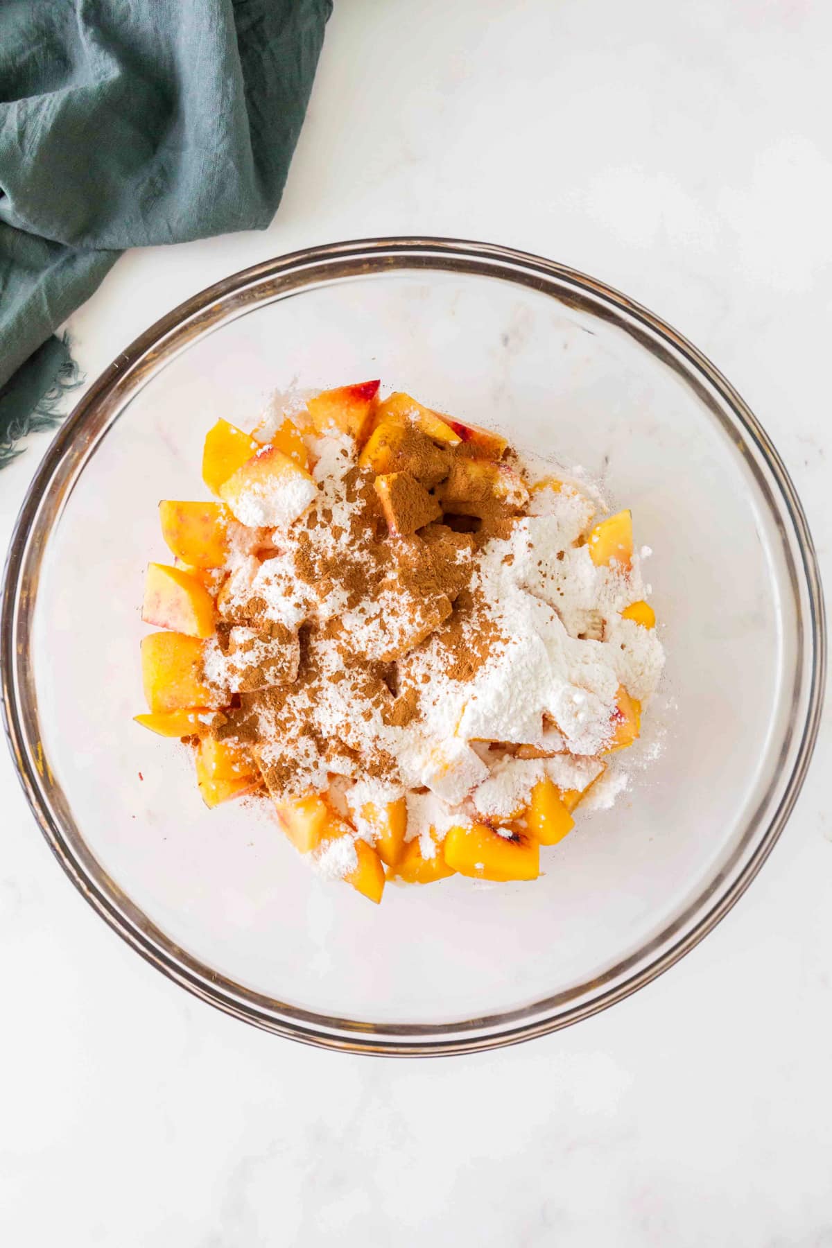 A glass bowl containing sliced peaches topped with sugar, flour, and cinnamon, on a white surface next to a dark green cloth. This is one of the processes in preparing Peach Pie.