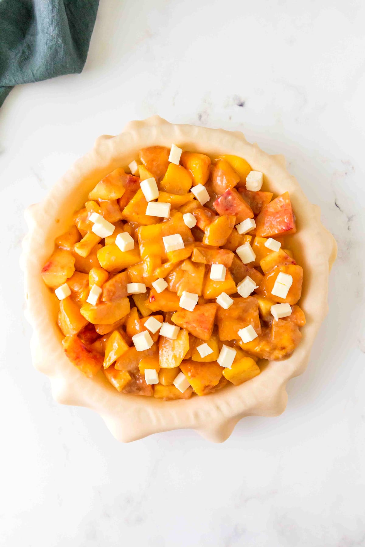 A pie crust filled with a mixture of diced peaches and small cubes of butter, ready to be baked. This is one process in preparing Peach Pie