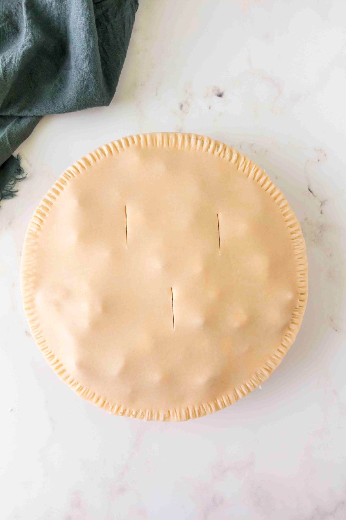 Unbaked Peach Pie with a crimped edge and three slits in the top crust, placed on a marble surface next to a green cloth.