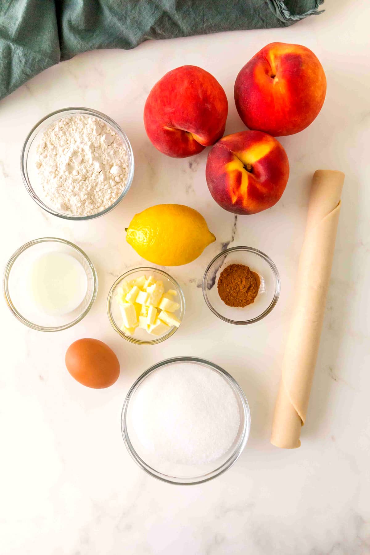 Ingredients for Peach Pie on a white counter include: homemade pie crust, peaches, granulated sugar, all-purpose flour, lemon juice, cinnamon, butter, egg and milk