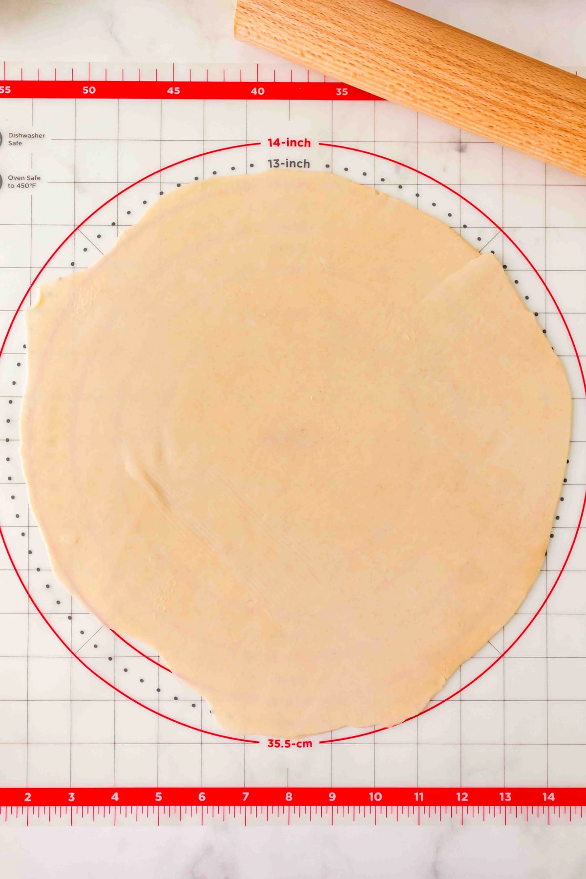 A rolled-out pie dough circle on a red and white pastry mat with measurements for Peach Pie recipe, next to a wooden rolling pin.