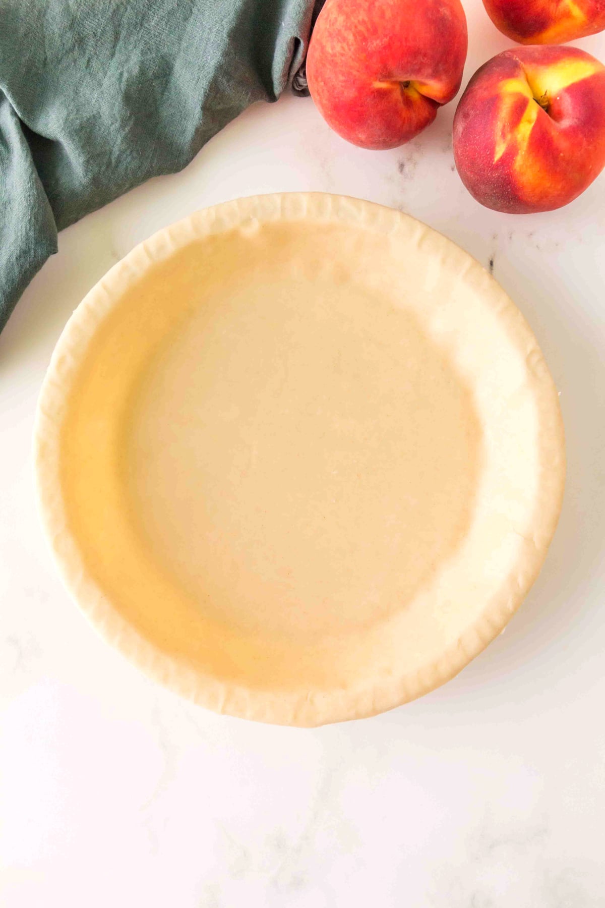 An empty pie crust in a baking dish sits on a white surface next to three whole peaches and a dark green cloth.