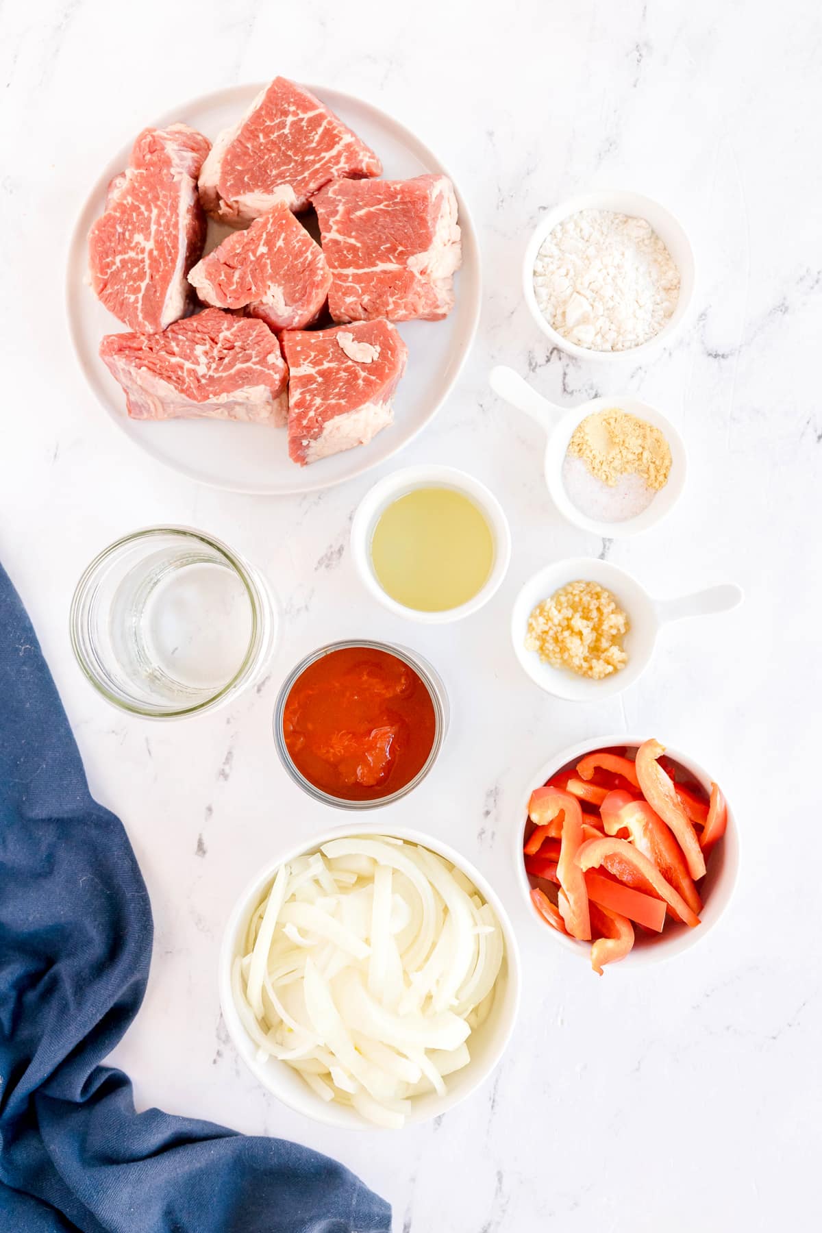 Ingredients for Swiss Steak are the following: boneless beef round, tipm or chuck steak, all purpose flour, ground mustard, salt, vegetable oil, whole canned tomatoes, garlic, water, onion, and bell pepper, arranged in bowls on a white surface.