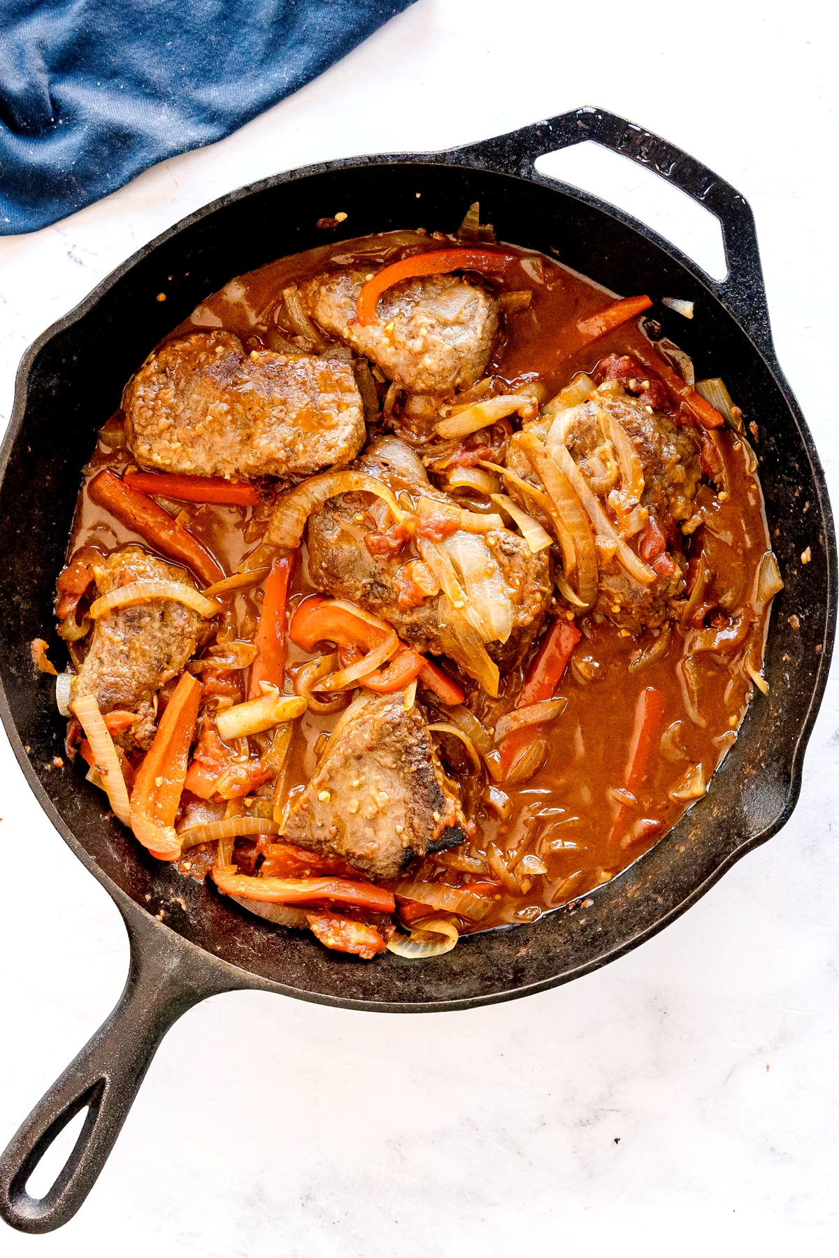 A cast iron of Swiss Steak, or a skillet filled with cooked beef steaks, sliced onions, and red bell peppers in a brown sauce.