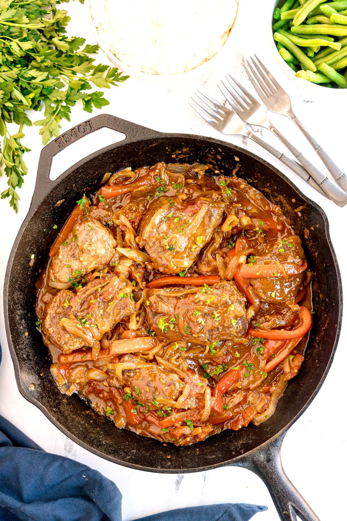 A cast iron skillet filled Swiss Steak or with cooked steak topped with sautéed onions, bell peppers, and sauce, garnished with chopped parsley.