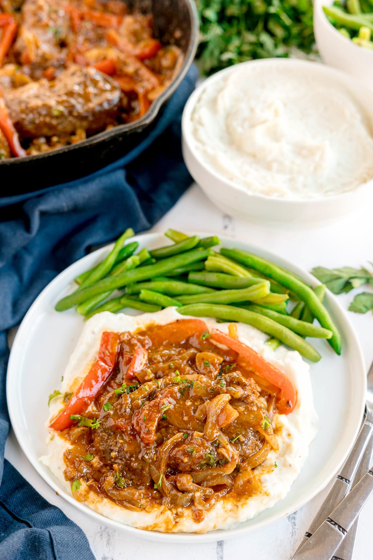 A plate of Swiss Steak with mashed potatoes topped with beef and vegetable stew, served with green beans. A bowl of mashed potatoes and a skillet of stew are in the background.