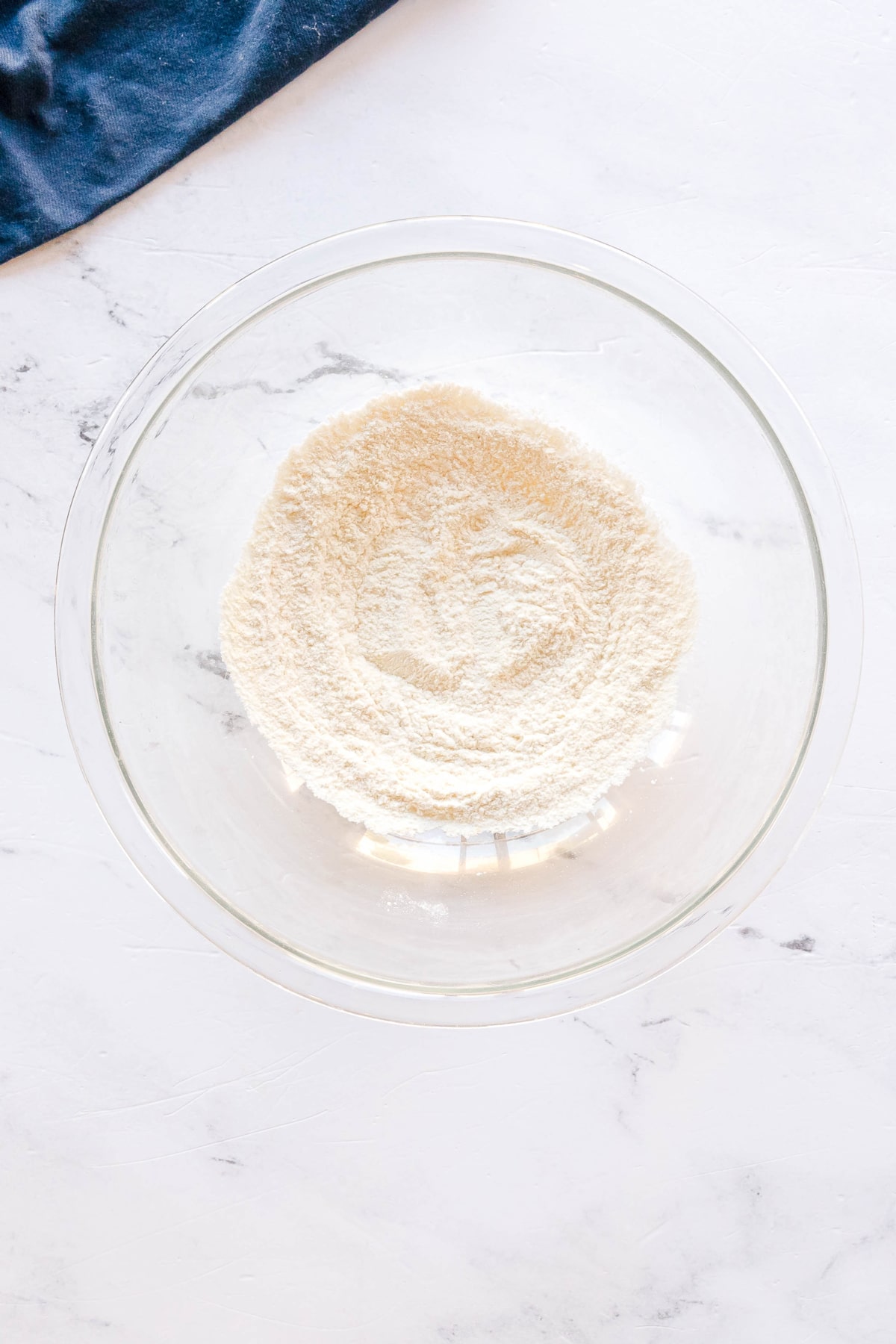 First step in preparing Swiss Steak is to use a glass bowl containing flour, ground mustard, and salt, on a white marble surface, with a corner of a blue cloth visible at the top left.