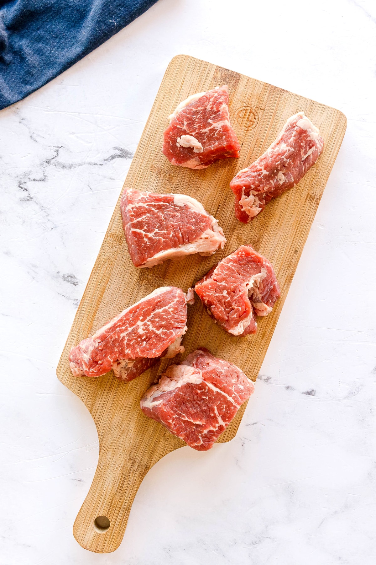 Six raw steak pieces are arranged on a wooden cutting board placed on a white marble surface for Swiss Steak Recipe.