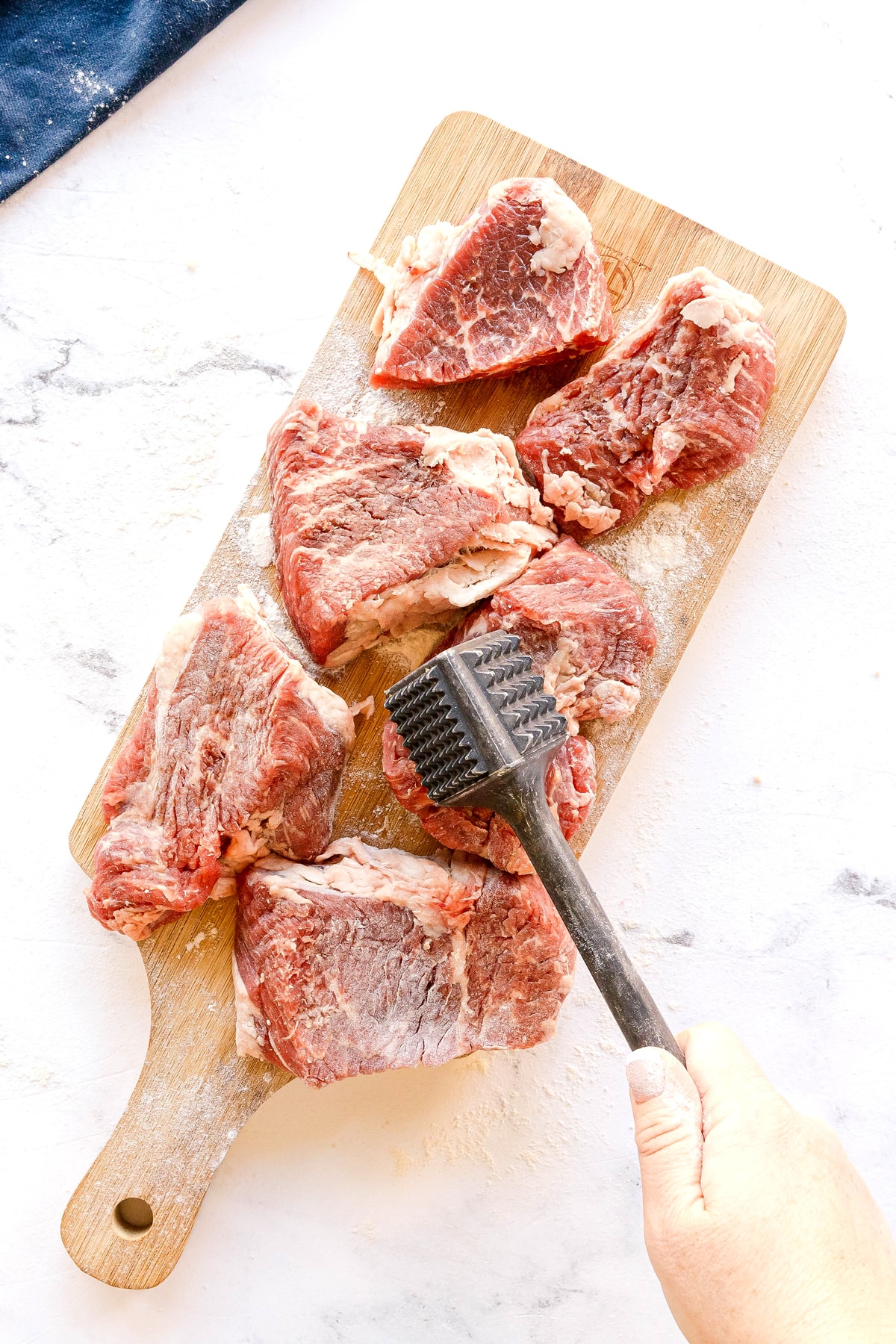 A hand uses a meat tenderizer to pound several raw beef pieces on a wooden cutting board for Swiss Steak recipe.