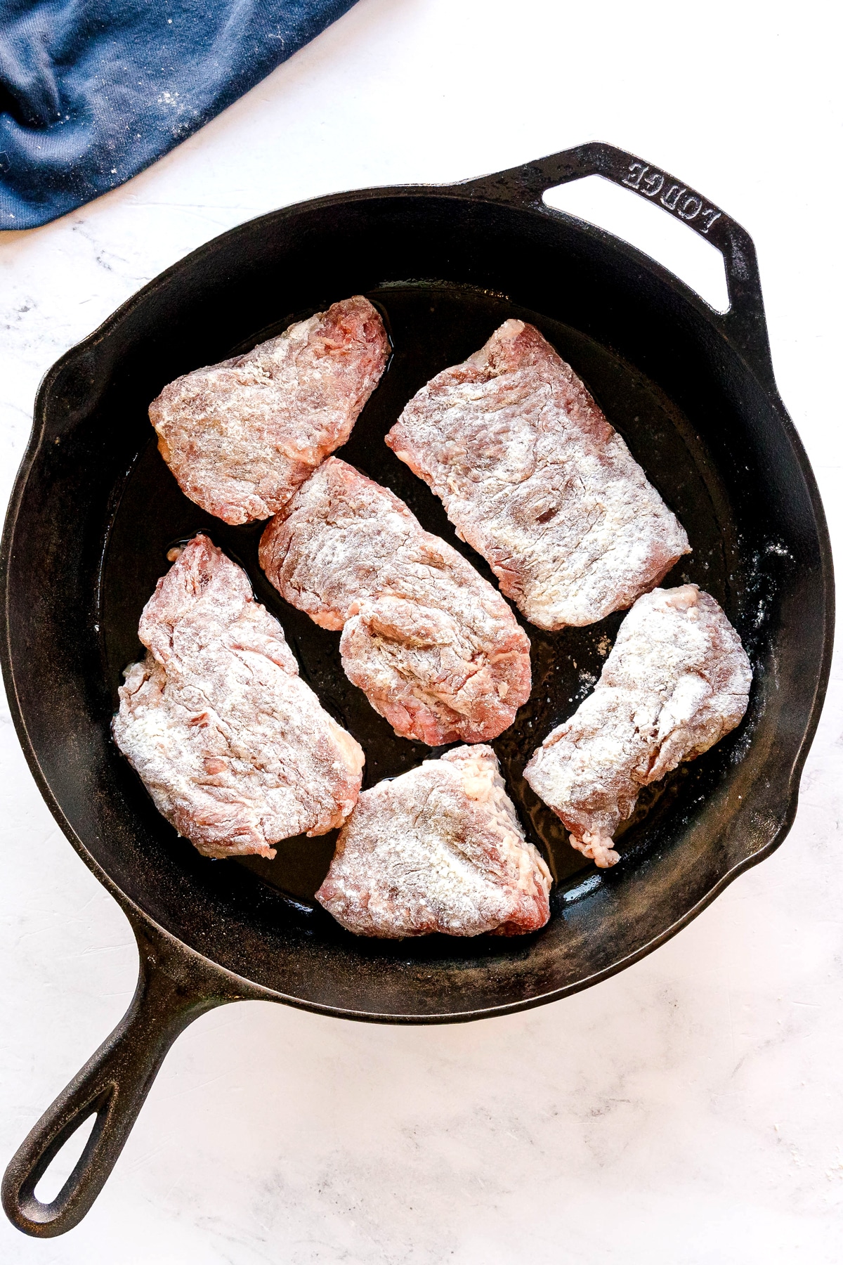 Six pieces of floured raw meat are arranged in a single layer inside a black cast iron skillet with vegetable oil on a white surface. This is one process in preparing Swiss Steak.