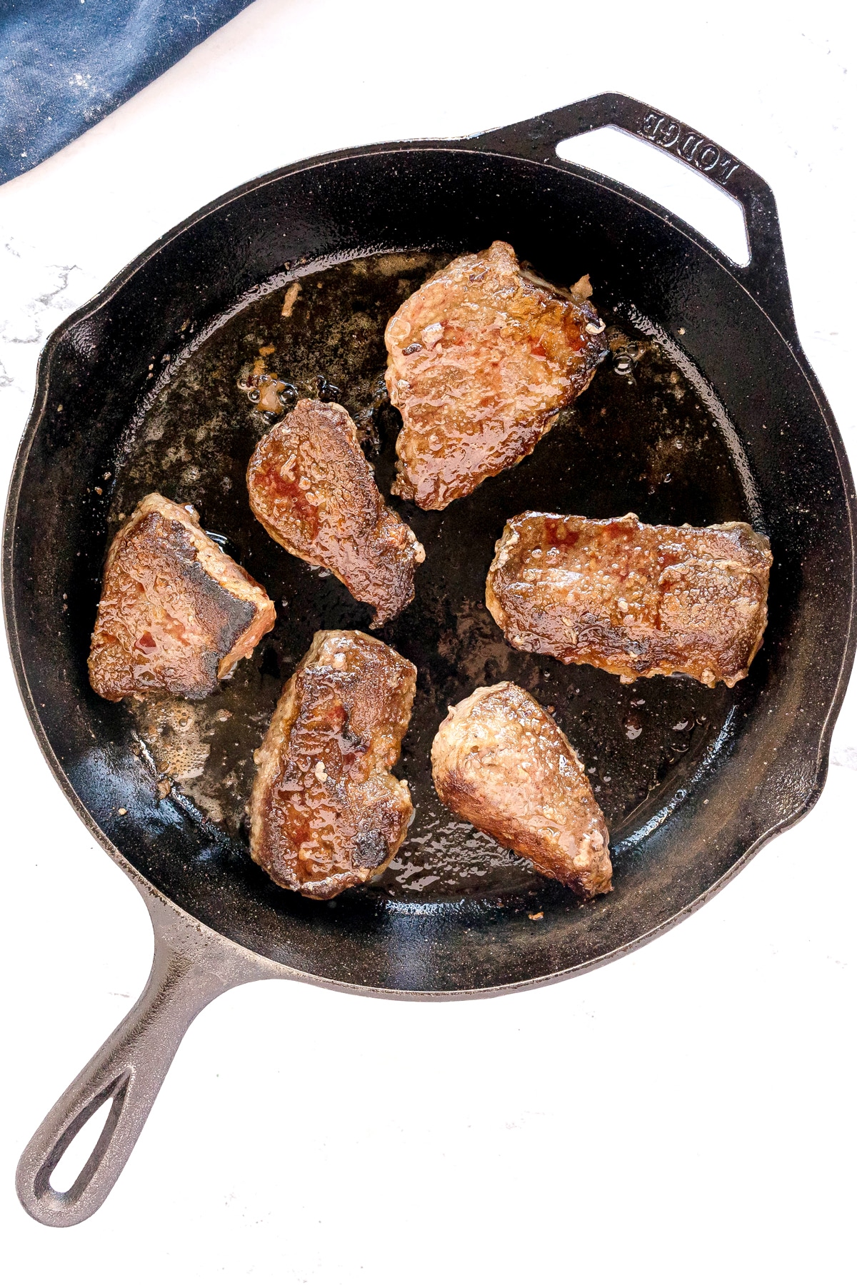 Six seared pieces of beef are cooking in a black cast iron skillet on a white surface, these are for Swiss Steak recipe.