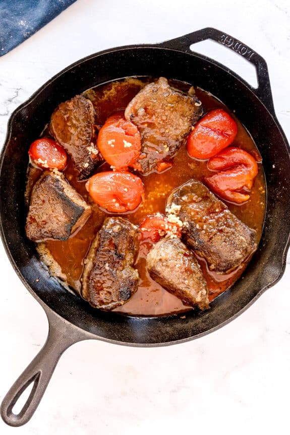 Next step in preparing Swiss Steak is to add garlic and tomatoes on a cast iron skillet containing seared pieces of beef, and a brown sauce on a light surface.