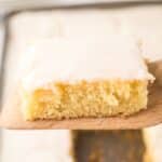 A square piece of yellow cake with white frosting on top, reminiscent of a classic white Texas sheet cake, rests on a wooden spatula.