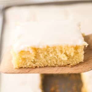 A square piece of yellow cake with white frosting on top, reminiscent of a classic white Texas sheet cake, rests on a wooden spatula.