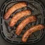 Four cooked sausages are arranged in a single layer inside an air fryer basket, making delicious Air Fryer Brats.