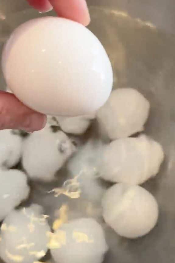 A hand holding a white egg over a pot of water with several eggs submerged inside.