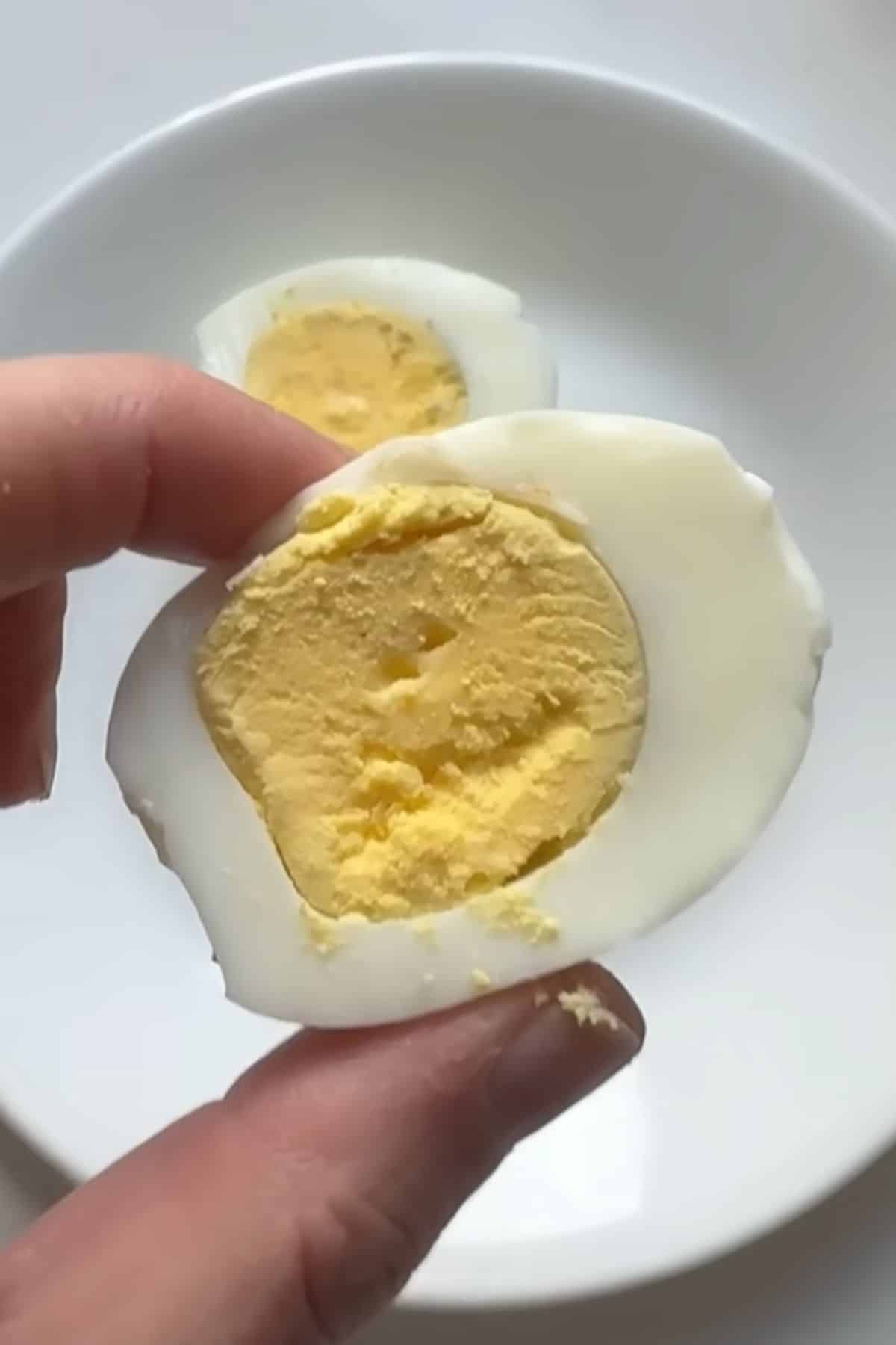 A hand holding a sliced hard-boiled egg with a pale yellow yolk over a white plate.