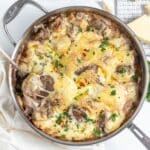 A pan of baked dish with melted cheese, browned on top, containing pieces of beef and garnished with chopped parsley. A serving spoon rests inside the pan.