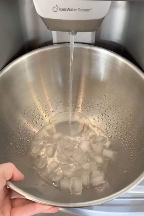 A metal bowl with ice cubes is being filled with water from a refrigerator dispenser.