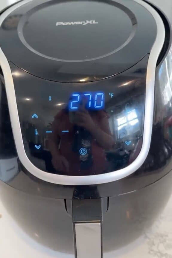 A digital air fryer displays a temperature of 270°F on its control panel, with the reflection of a person visible on the surface.