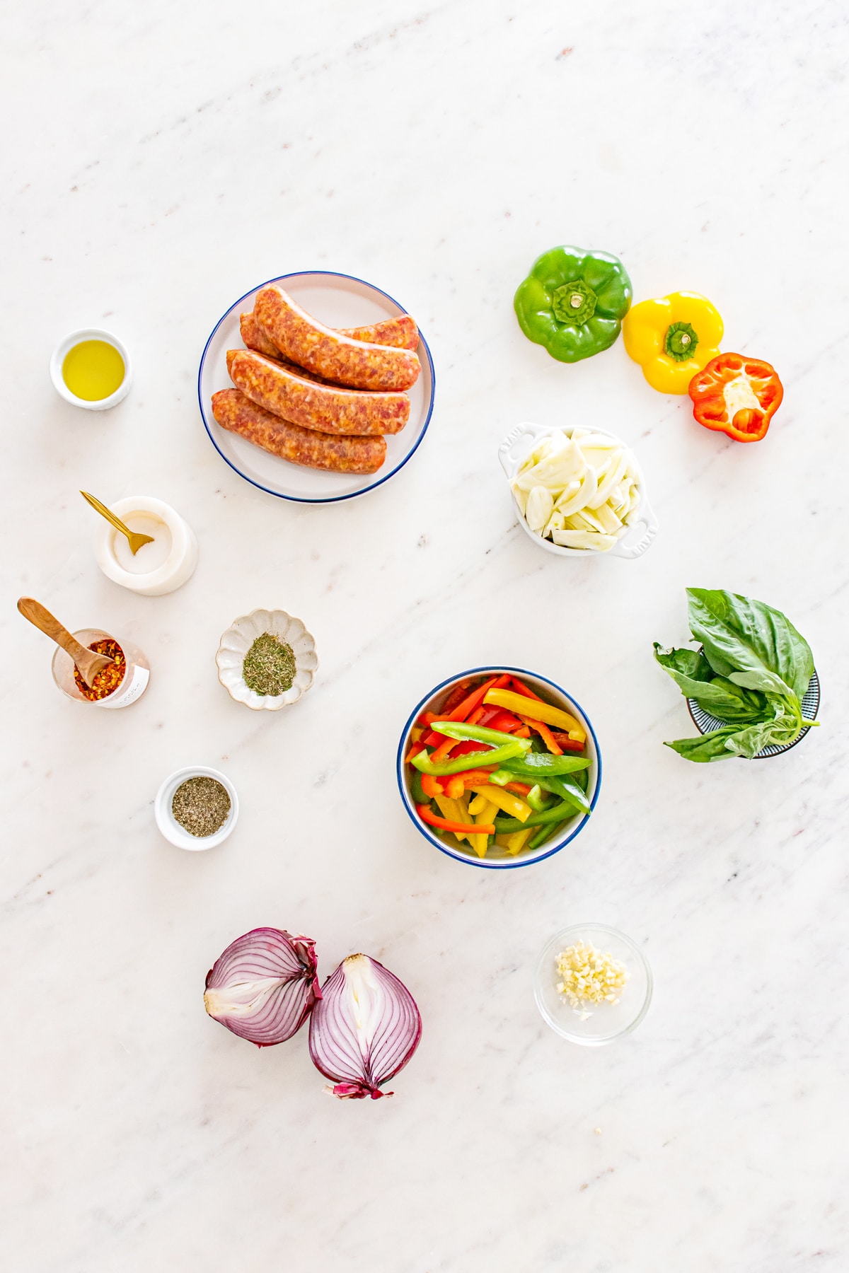 Ingredients for Sheet Pan Sausage and Peppers include: A flat lay of hot Italian sausages, sliced bell peppers, red onion, garlic, fennel, fresh basil, olive oil, kosher salt, black pepper, italinan seasoning, and crushed red pepper arranged on a white marble surface.