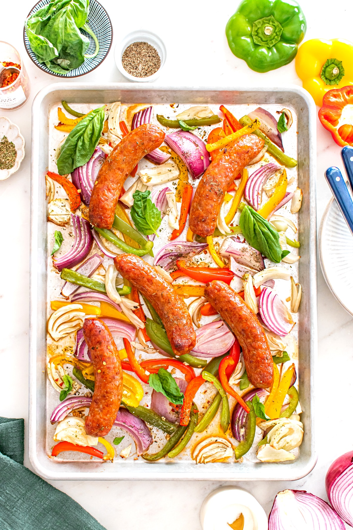 A tray of Sheet Pan Sausage and Peppers fresh from the oven. Sliced red onions, bell peppers, and herbs, arranged on a white surface with fresh vegetables and small bowls can be seen nearby.