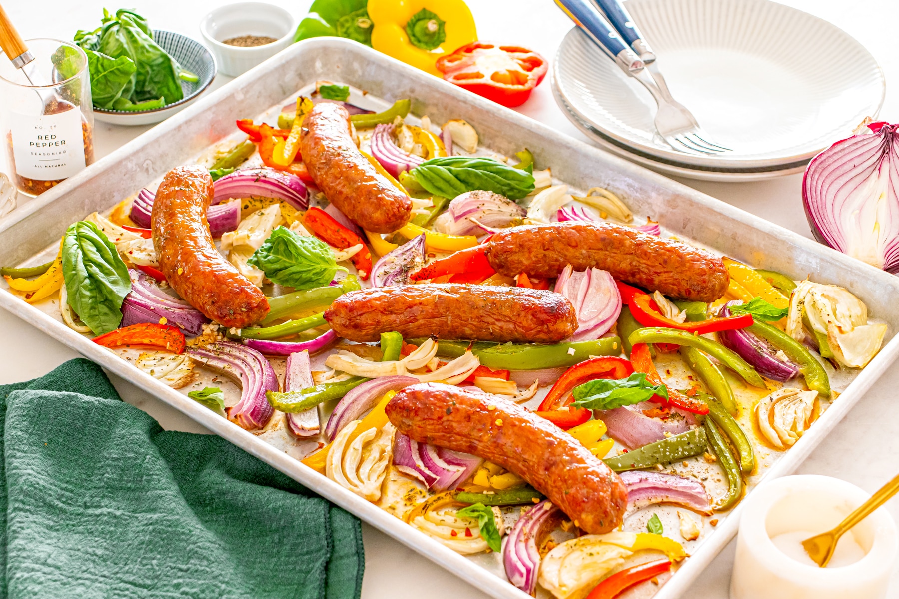Sheet Pan Sausage and Peppers with cooked sausages, sliced red onions, bell peppers, and basil leaves, surrounded by plates, utensils, and condiments on a white countertop.