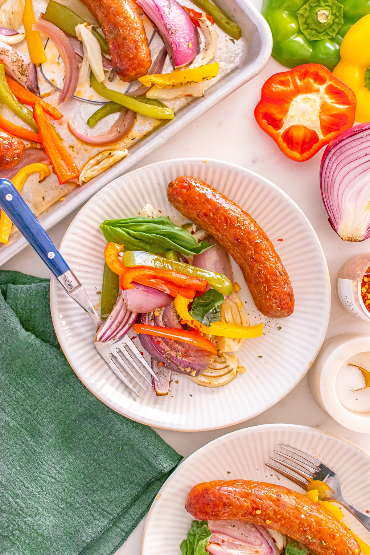 Two plates of Sheet Pan Sausage and Peppers with sausages, roasted bell peppers, and onions, next to a sheet pan with more vegetables and sausages, and fresh sliced peppers and onion on the table.