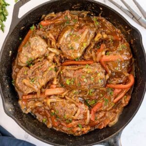 A cast iron skillet filled with cooked steak pieces, bell peppers, onions, and tomato-based sauce—this delicious Recipe For Swiss Steak is garnished with chopped parsley.