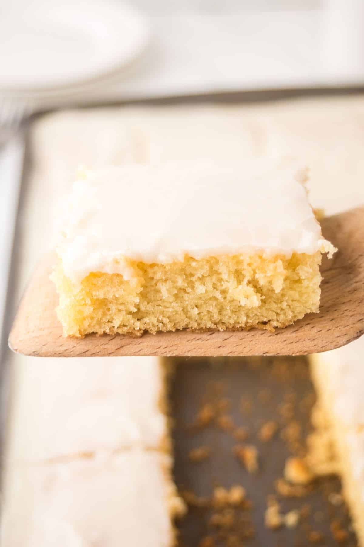 A square piece of yellow cake with white icing, reminiscent of a white Texas sheet cake, is held up on a wooden spatula above a pan of similar cake pieces.