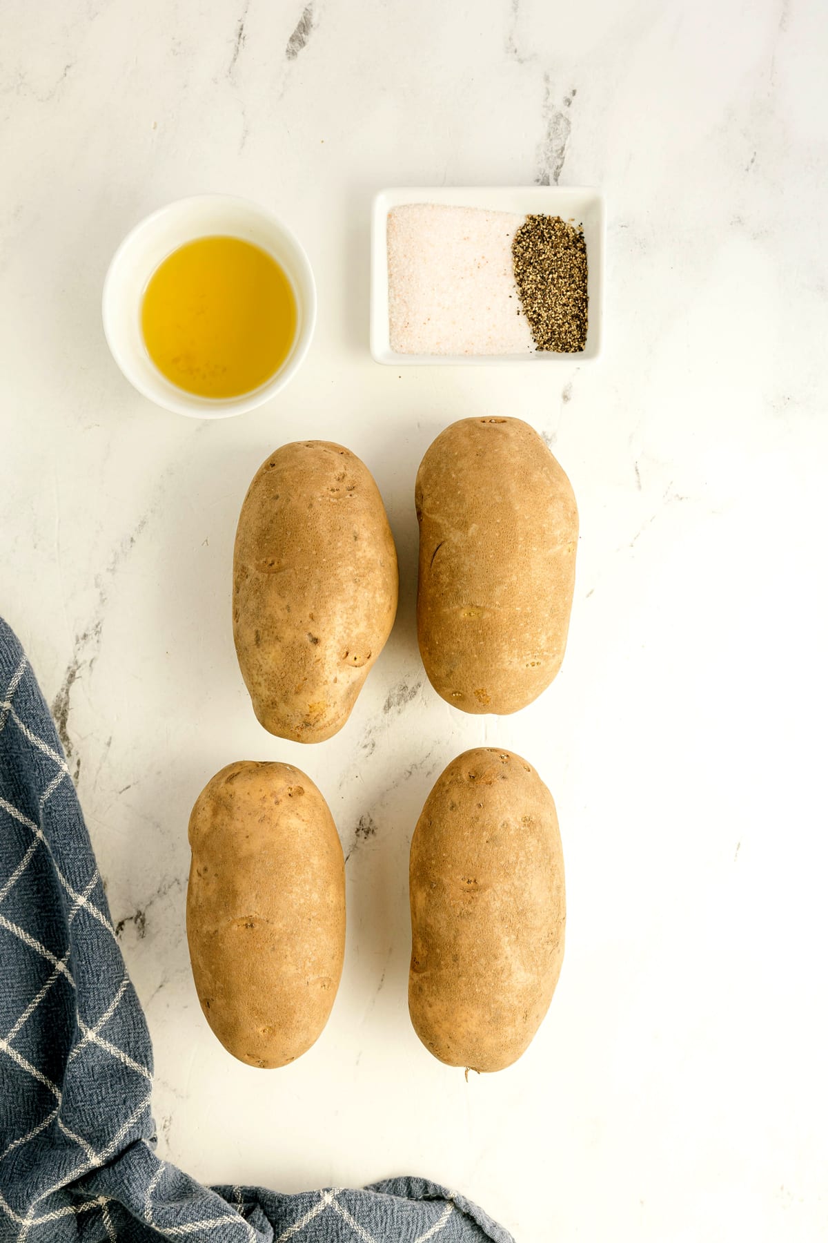 Ingredients for Air Fryer Baked Potato recipe include four raw potatoes with a small bowl of olive or avocado oil, a dish of salt and pepper, and a blue checkered towel on a white surface.