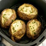 Four baked potatoes with crispy skins, each topped with a pat of butter and sprinkled with chopped parsley, inside an air fryer basket.