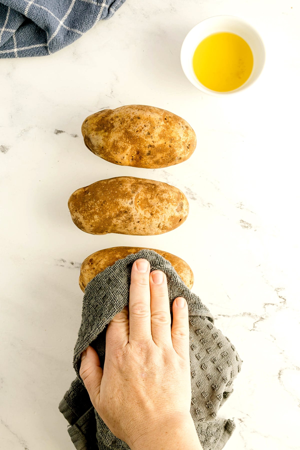 A hand uses a gray cloth to clean one of three raw potatoes on a white countertop, with a small bowl of oil nearby. This is the first step in preparing Air Fryer Baked Potato recipe