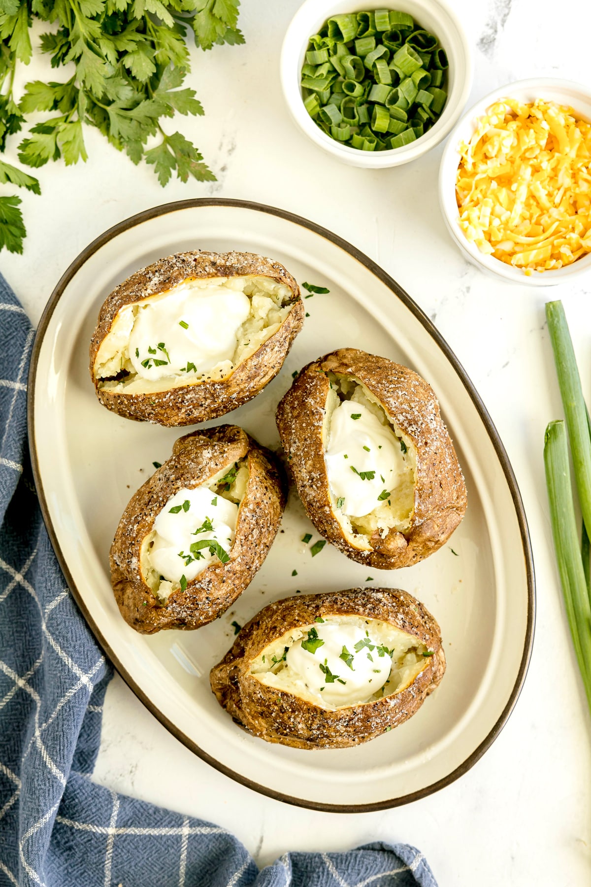 Four baked potatoes topped with sour cream and chopped parsley on a white oval plate, with bowls of chopped green onions, shredded cheese, and fresh herbs nearby.