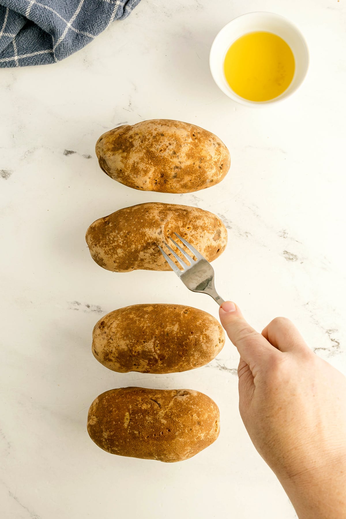 Next step in preparing Air Fryer Baked Potato is to gather four raw potatoes on a white surface, a hand piercing one with a fork, a bowl of oil, and a blue cloth visible in the background.