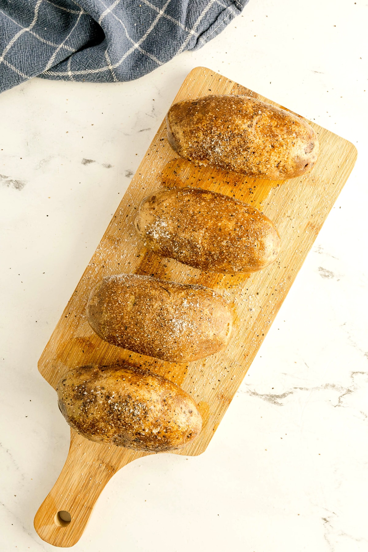 Four seasoned baked potatoes arranged in a row on a wooden cutting board, with a blue checkered towel nearby on a white surface. This is another step in preparing Air Fryer Baked Potato.