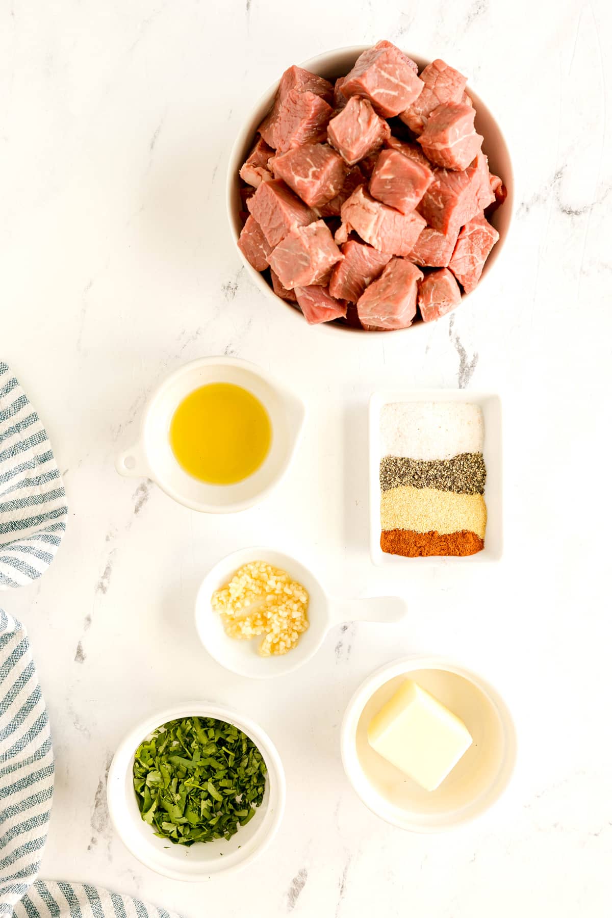 Overhead view of ingredients for Air Fryer Steak Bites include sirloin steak in a bowl, with small bowls of olive oil, kosher salt, black pepper, garlic powder, smoked paprika, butter, garlic cloves, and fresh parsley on a white counter.