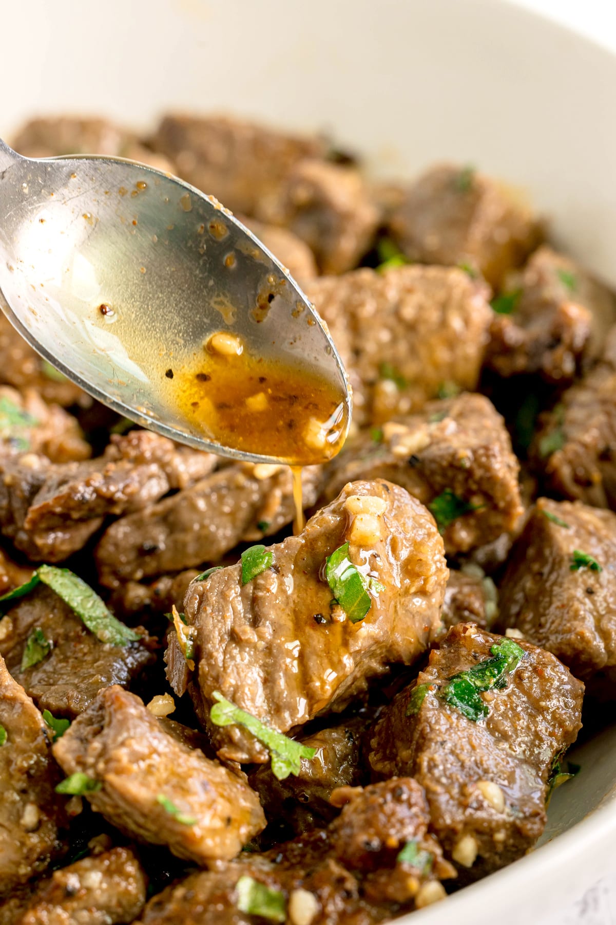 A spoon drizzles sauce over chunks of cooked Air Fryer Steak Bites garnished with chopped herbs in a white dish.
