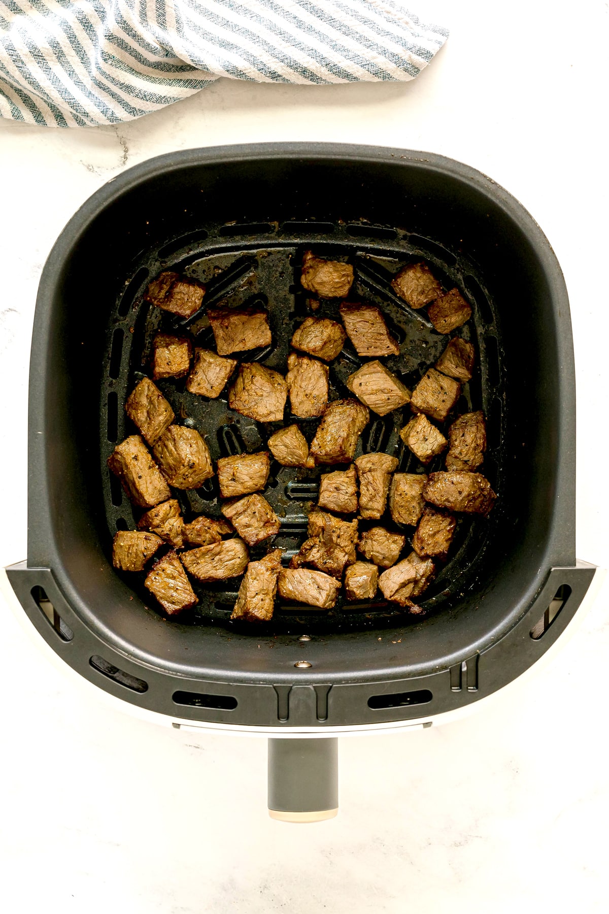 Cubed pieces of cooked steak for Air Fryer Steak Bites recipe, arranged in a single layer inside an air fryer basket on a white surface.