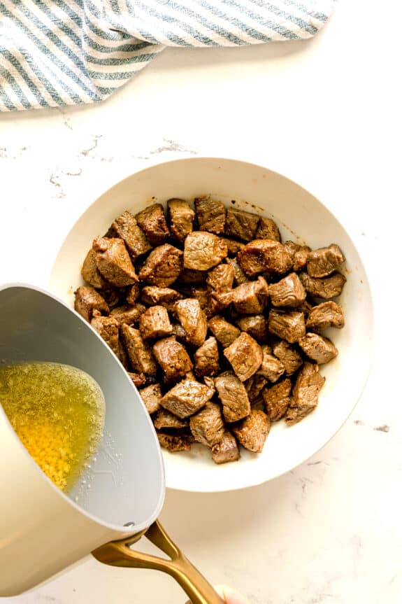 A pot of melted butter and with minced garlic is being poured over a bowl filled with cooked beef cubes on a white surface. This is one of the steps in preparing Air Fryer Steak Bites