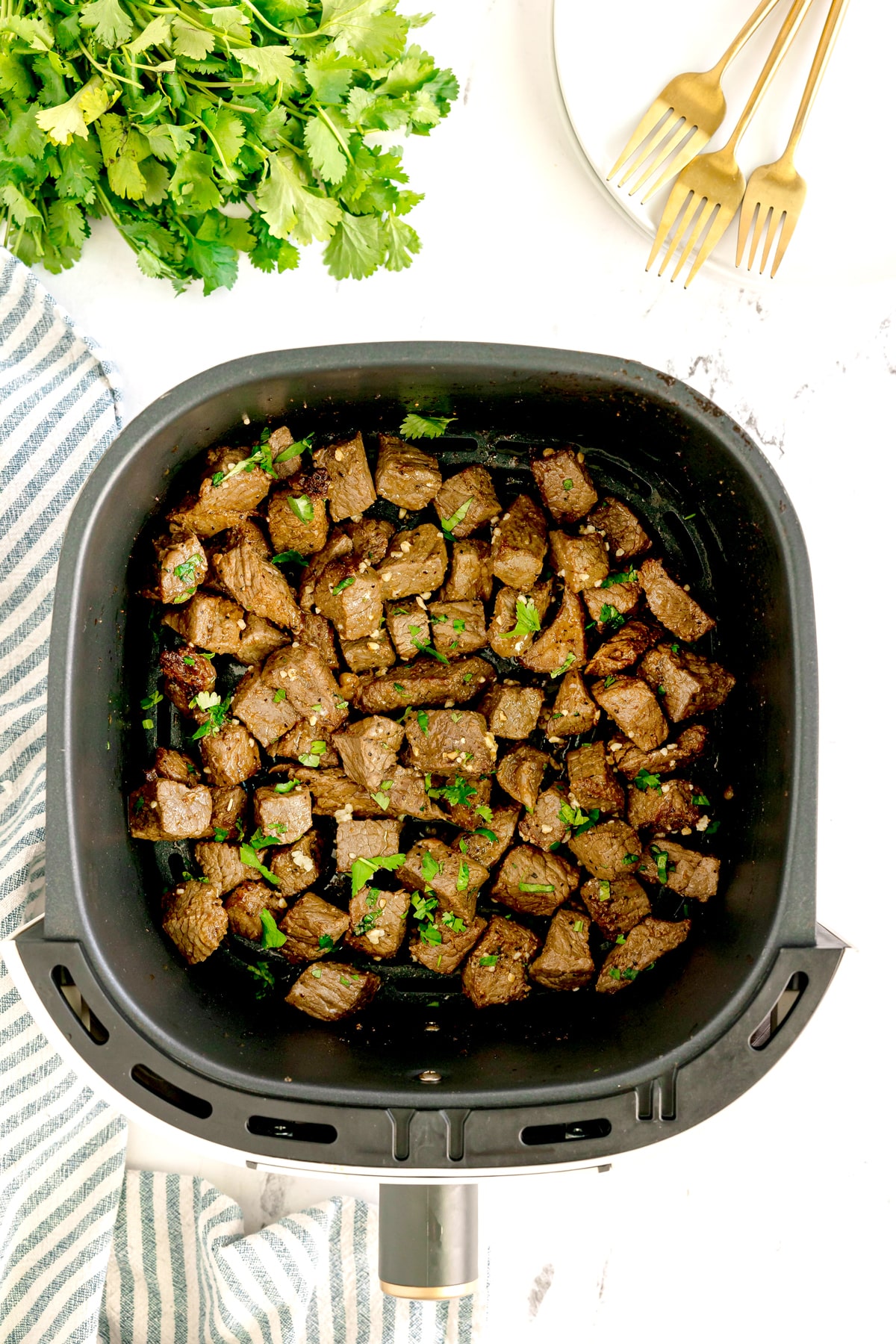 Air Fryer Steak Bites garnished with herbs in an air fryer basket, with parsley, a striped towel, and plates with gold forks nearby.