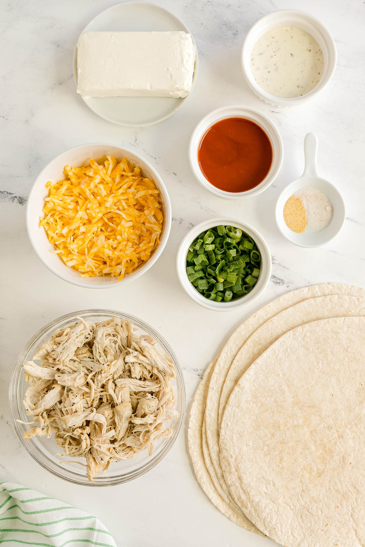 Ingredients for buffalo chicken pinwheels laid out, including shredded chicken, flour tortillas, shredded cheese, cream cheese, chopped green onions, ranch dressing, hot sauce, and seasonings in bowls.